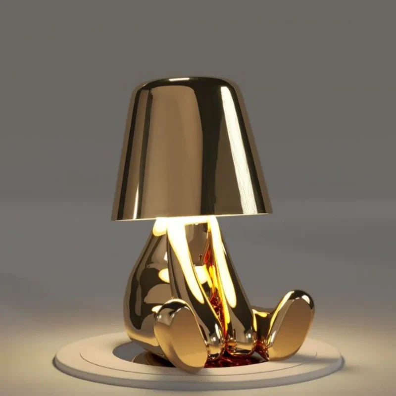 Designerska lampka nocna z postacią siedzącą i metalicznym połyskiem w ciepłym świetle
