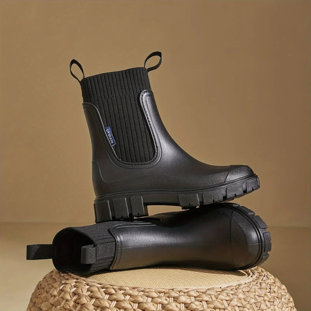 Women's Chelsea boots in black with ribbed elastic gussets and chunky tread sole displayed on woven base